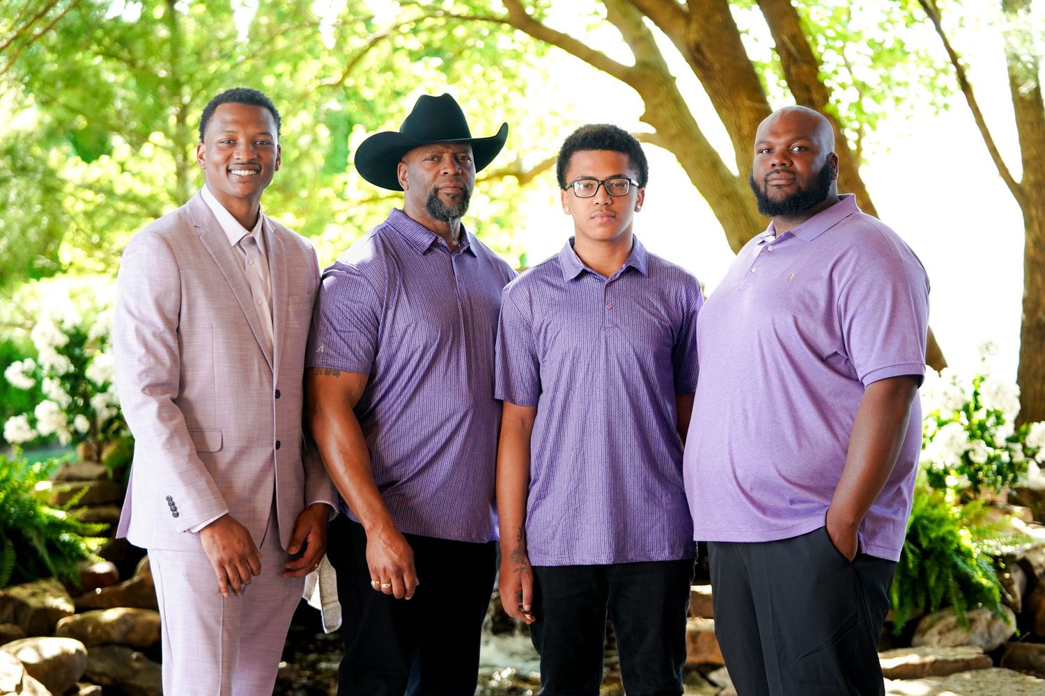 A group of groomsmen in matching purple shirts and black pants pose together at an outdoor wedding venue.