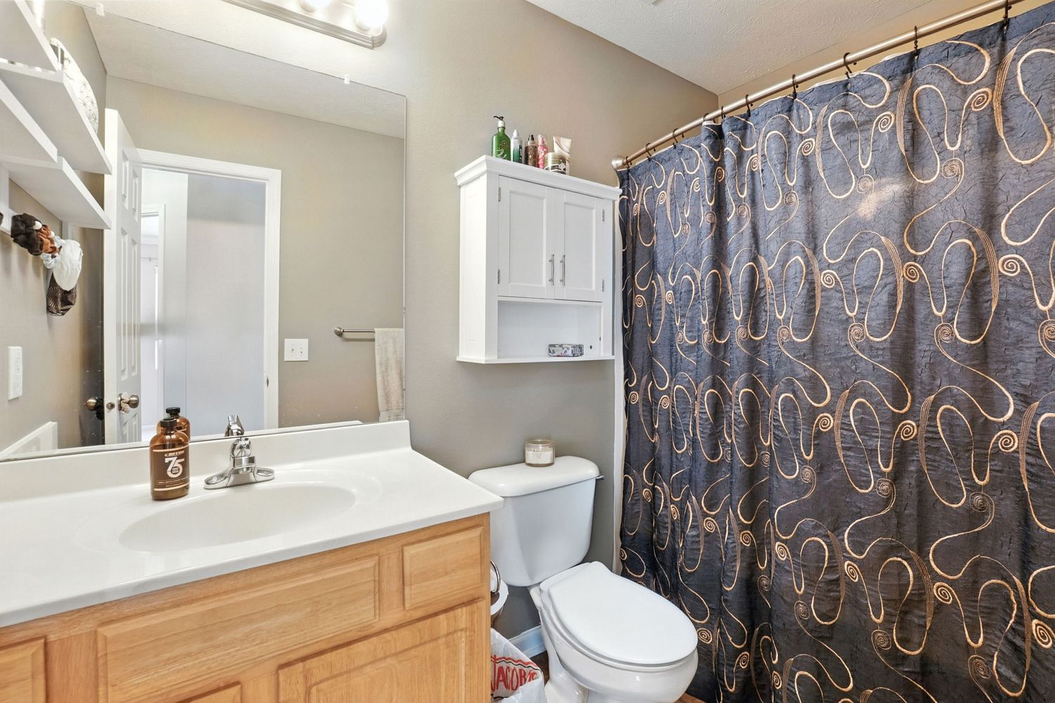 Bathroom with oak vanity, decorative shower curtain and white storage cabinet above toilet.