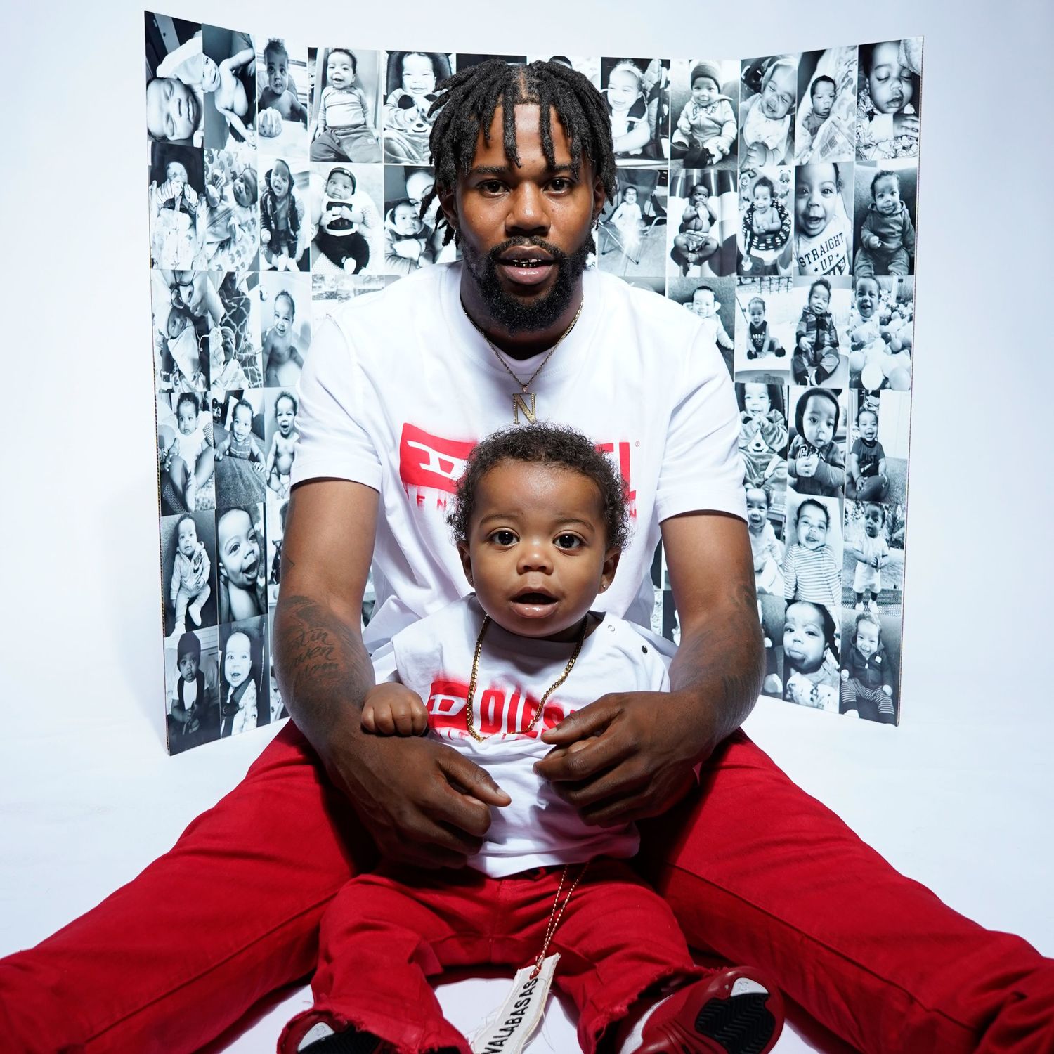 A person in a white t-shirt and red pants sits with a small child against a wall decorated with black and white photos.