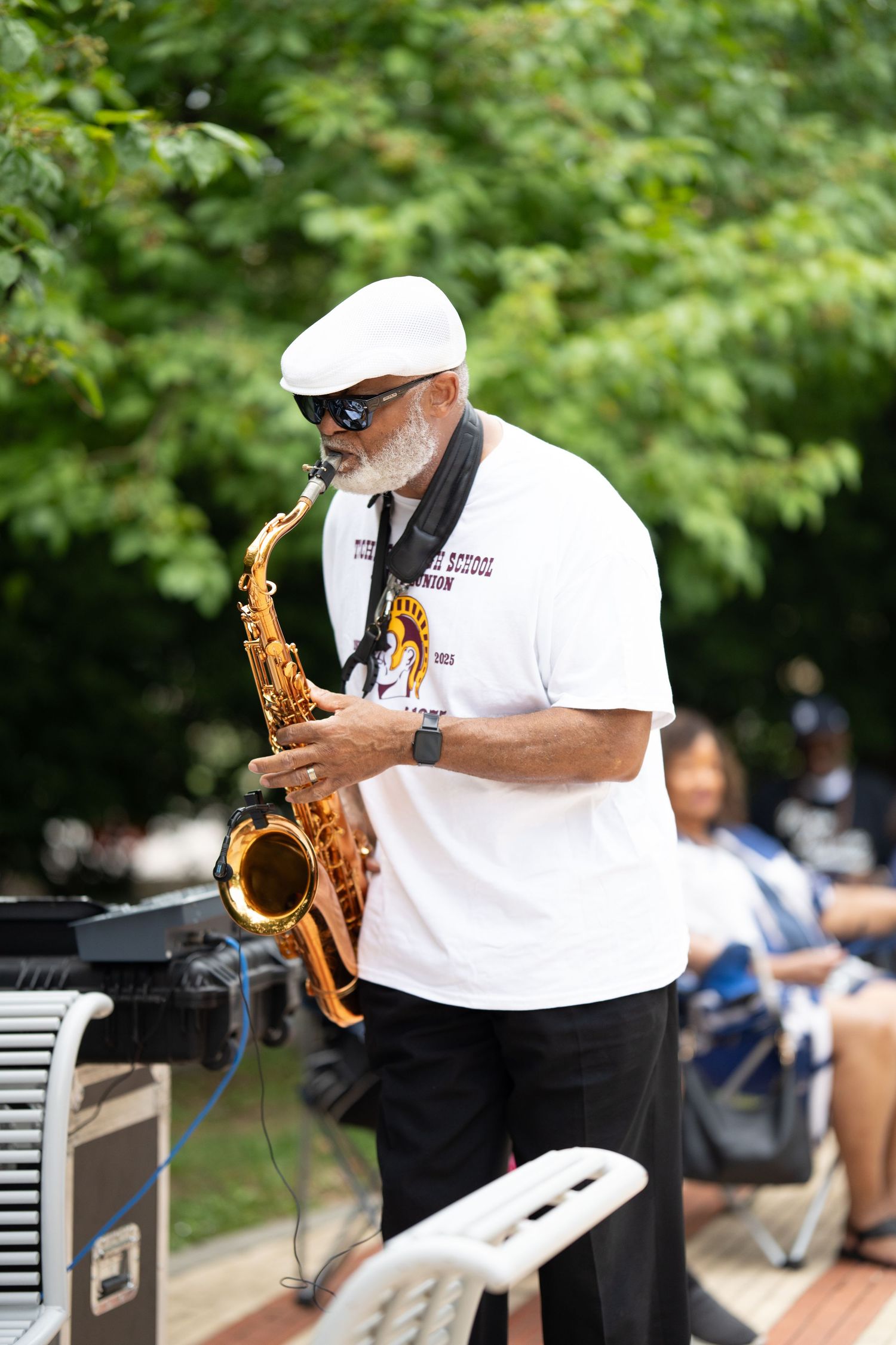 Musician playing saxophone at outdoor performance wearing white shirt and cap among green trees.