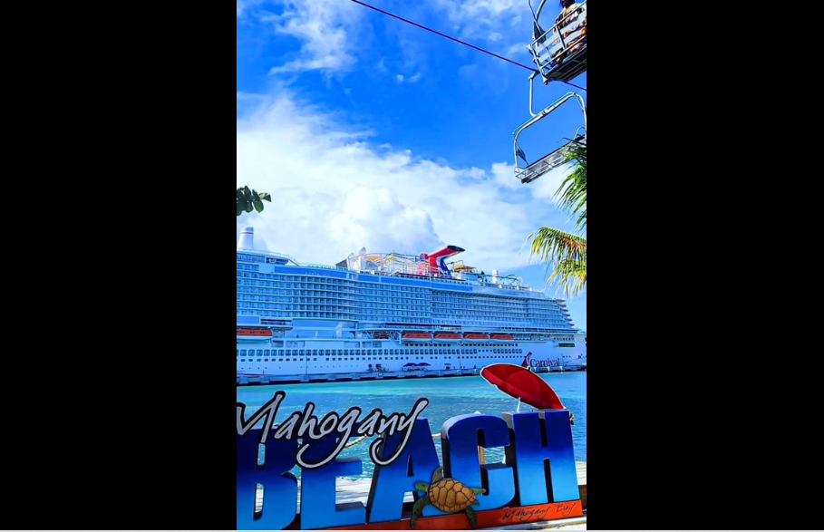 A Carnival cruise ship docked at port with its distinctive red funnel visible against a bright blue sky with palm trees.