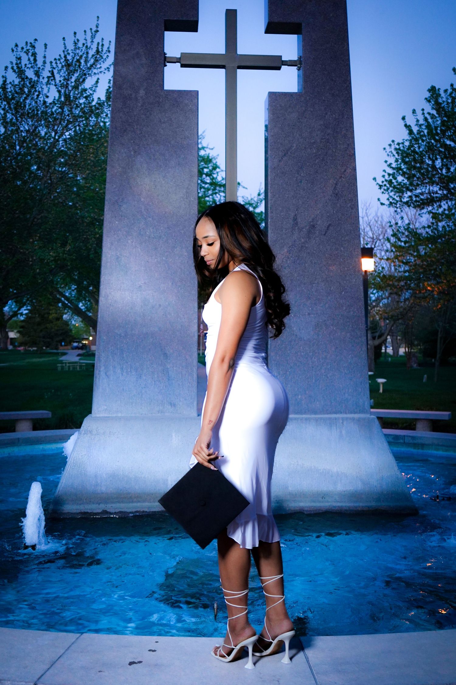 Graduate in white dress poses near architectural columns at dusk.