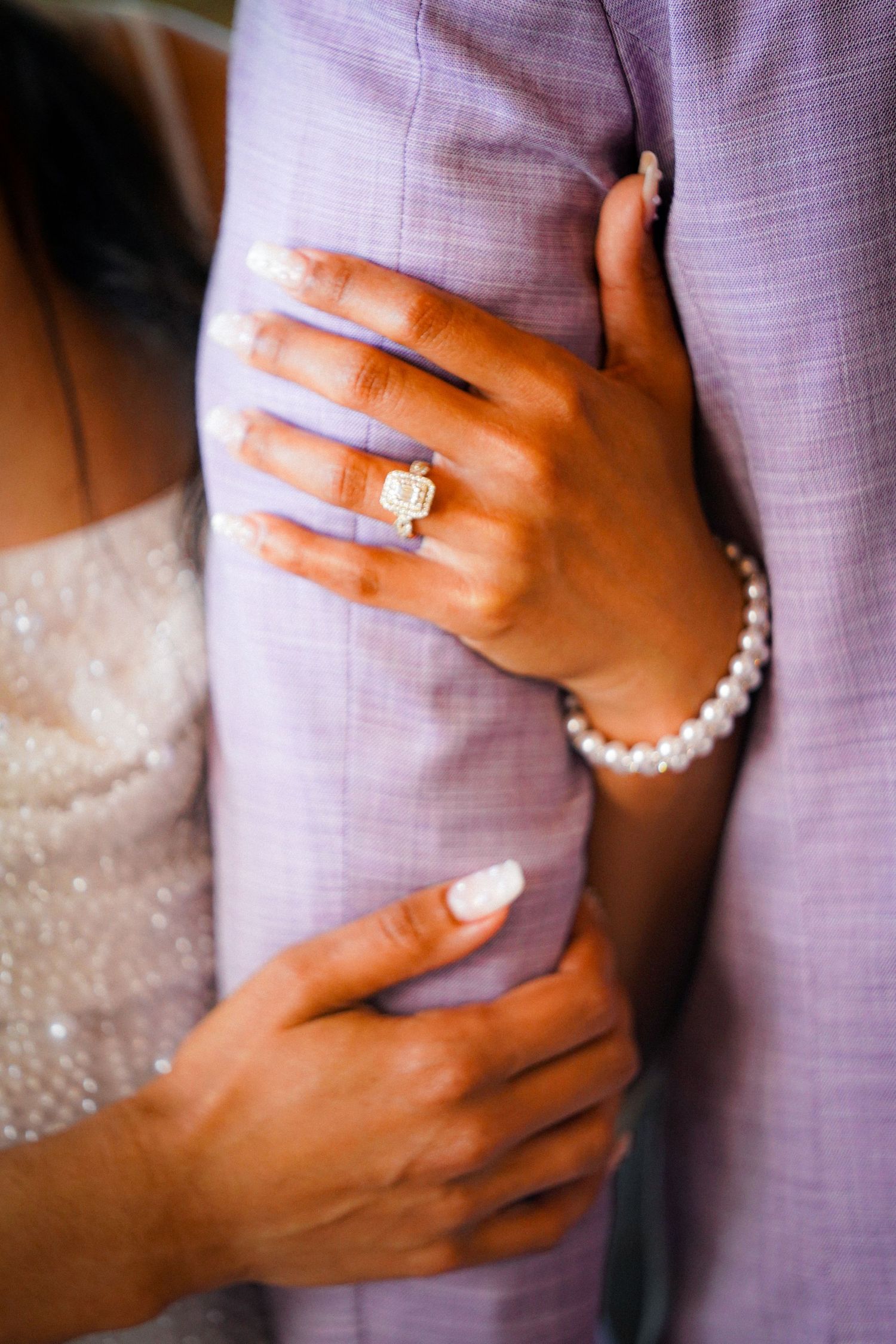 Close-up of hands wearing pearl bracelet and engagement ring against a light purple fabric background.