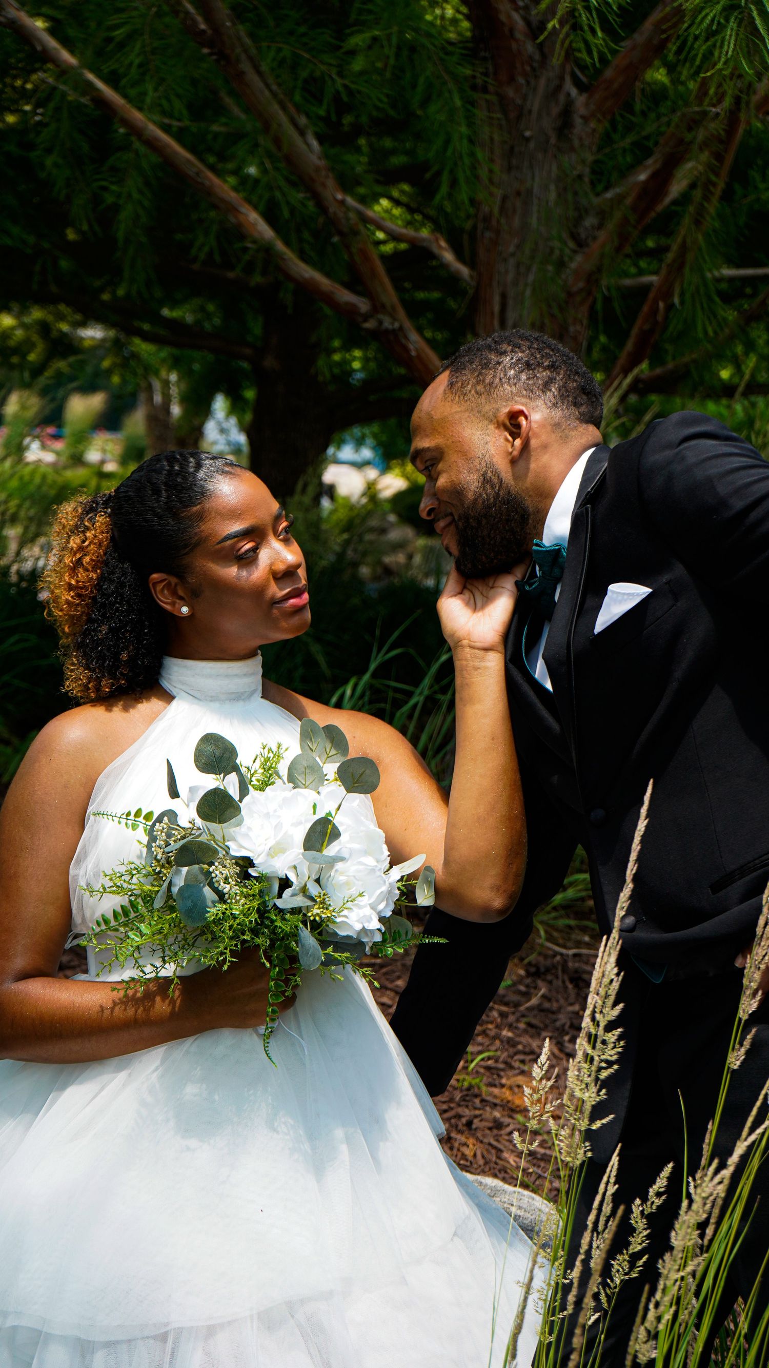 A romantic wedding couple shares intimate moments in a garden setting with natural greenery and white floral details.