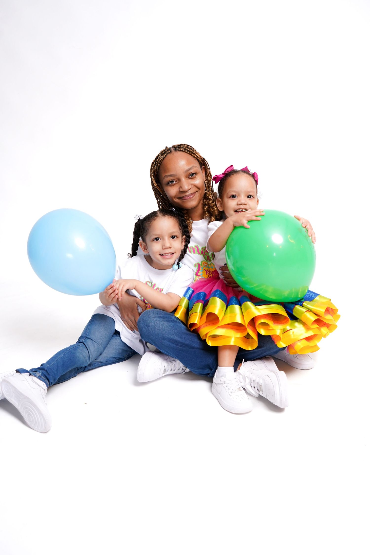 Three children in colorful outfits sit together holding blue and green balloons against a white background.