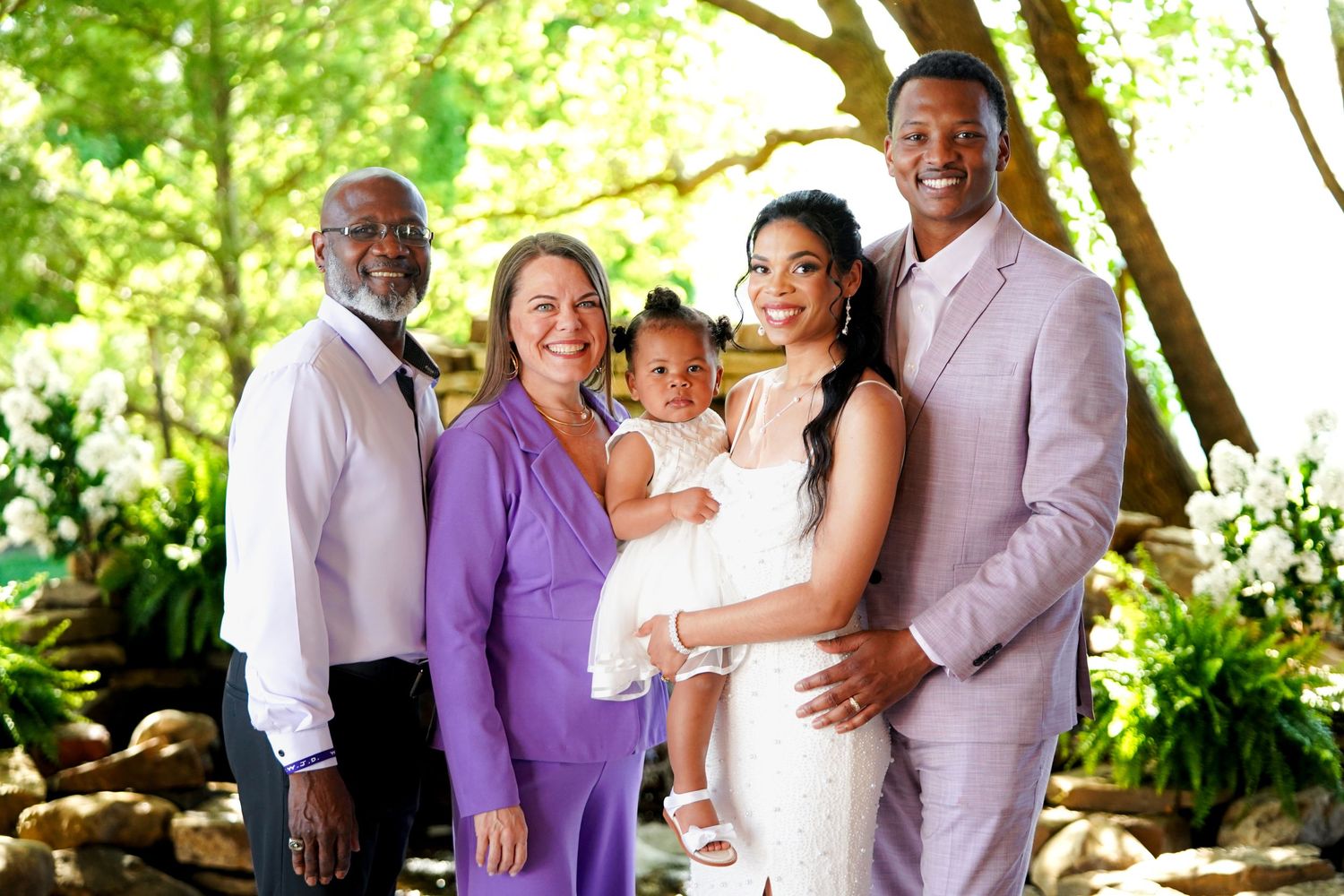 Wedding party members in purple and white attire share joyful embraces outdoors under trees.