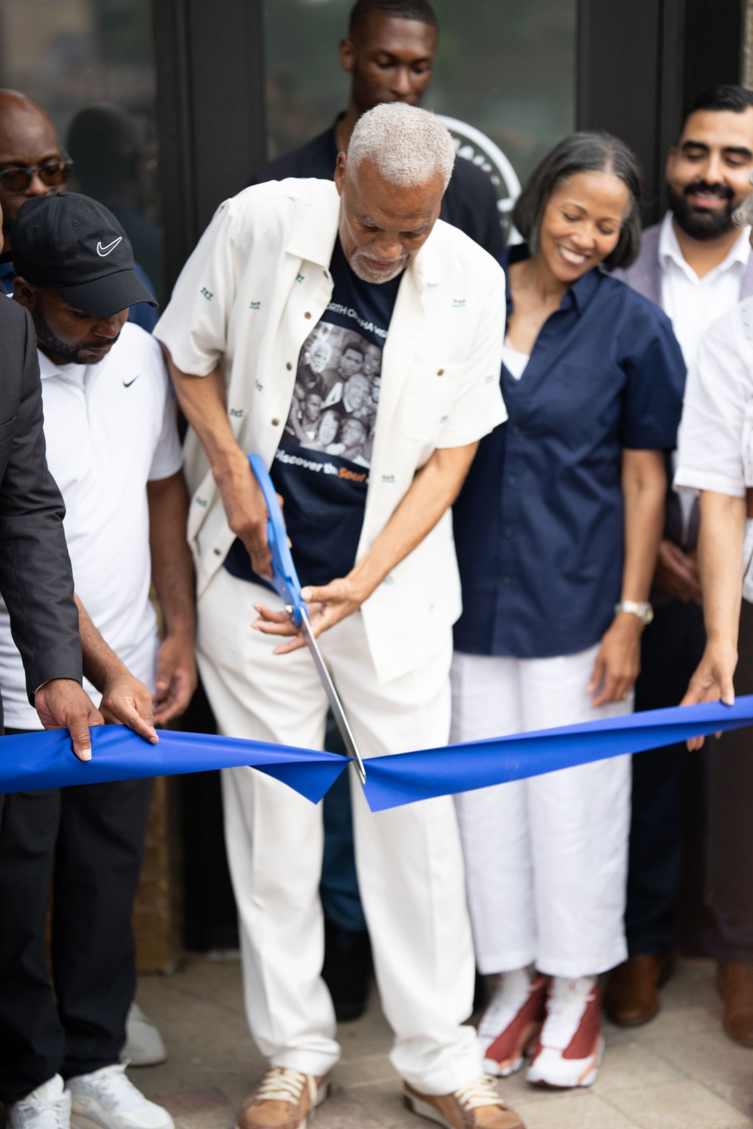 A group of people in white and navy attire cut a ceremonial blue ribbon at an opening event.