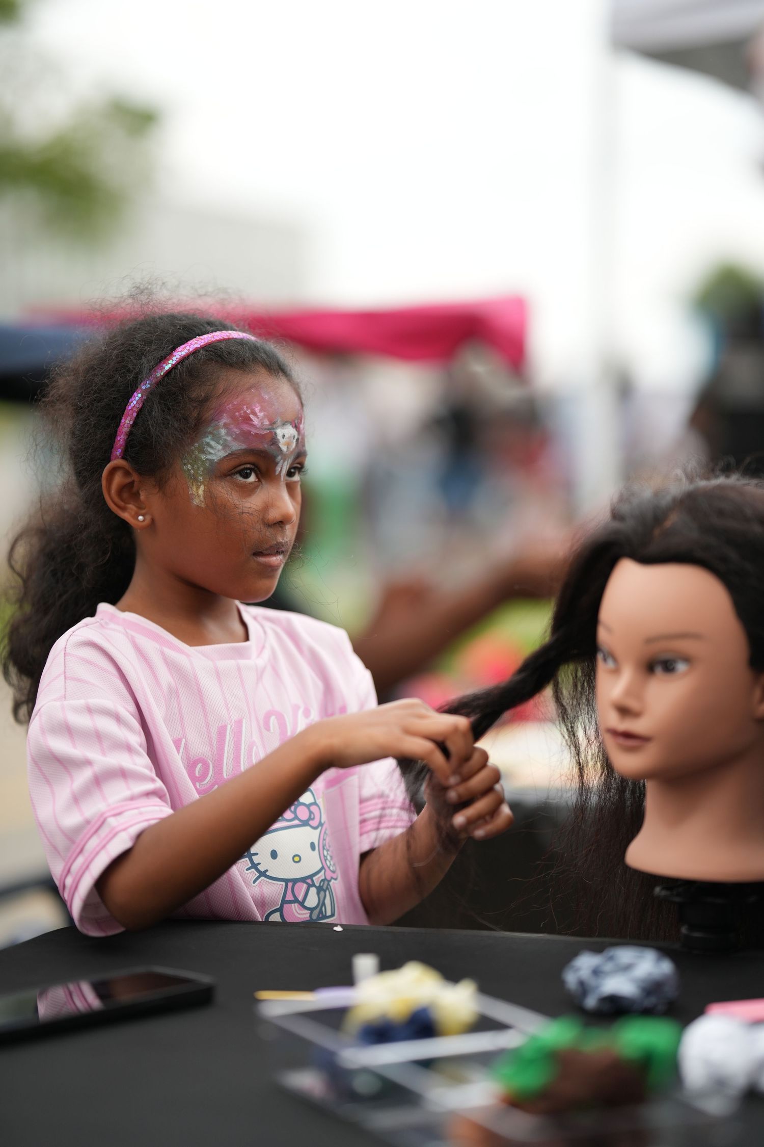 A child wearing pink practices face painting techniques on a mannequin head during an outdoor event.