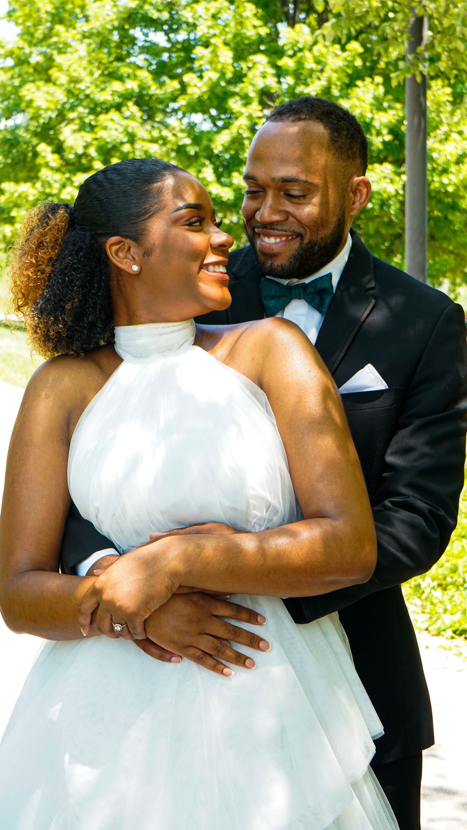 A joyful wedding couple embraces and laughs together outdoors with lush green foliage in the background.