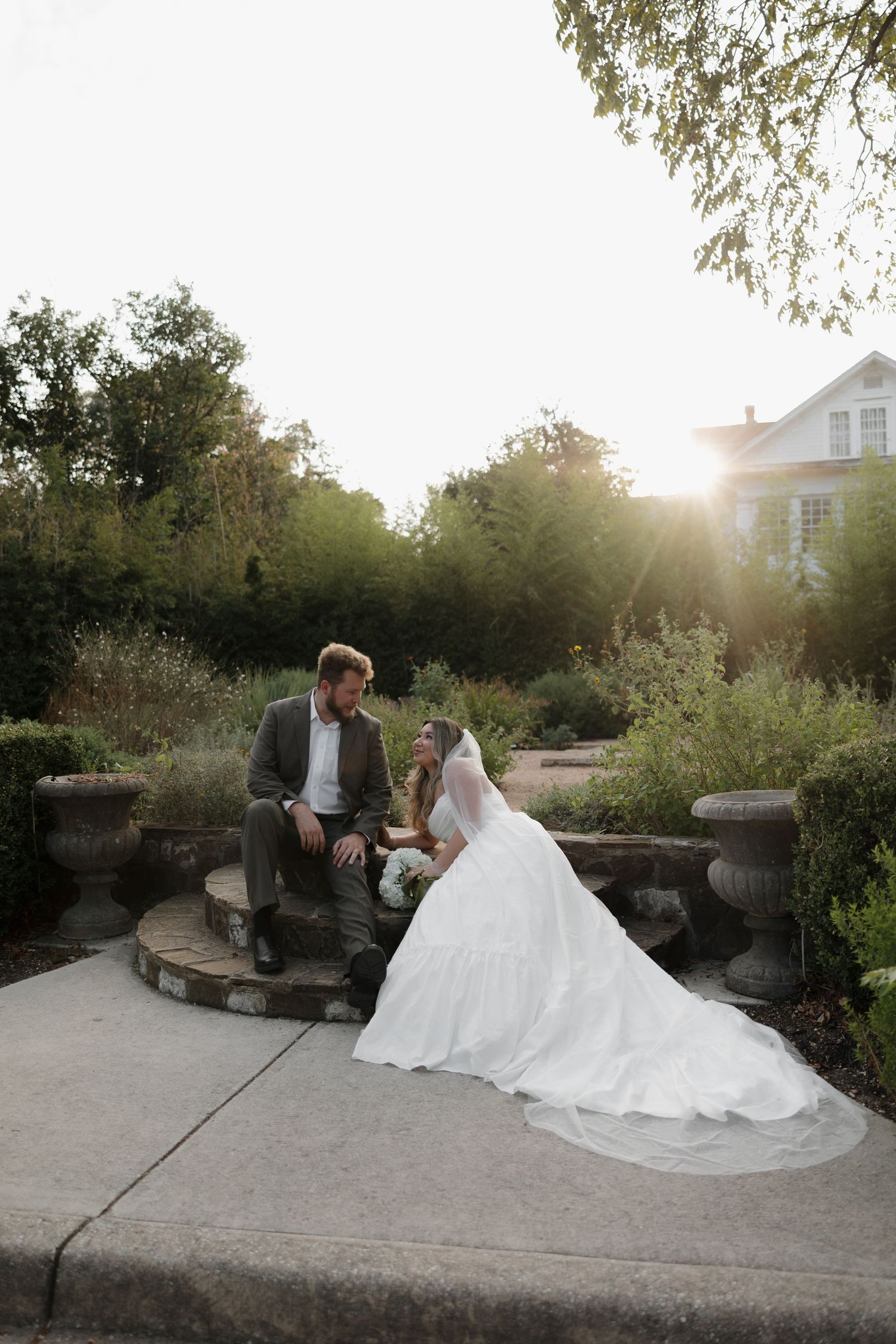 Newlyweds share a quiet moment on stone steps during sunset in a garden.