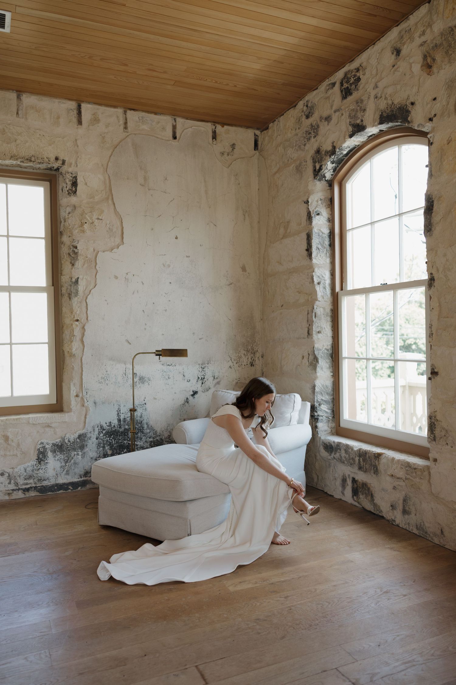 A figure in white fabric lies gracefully on a wooden floor in an empty room with distressed walls and tall windows.