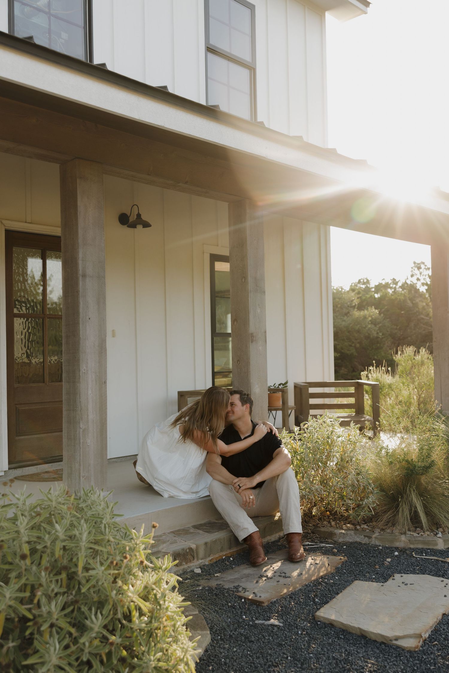 A couple sits together on the front steps of a modern farmhouse porch at sunset, surrounded by landscaping.