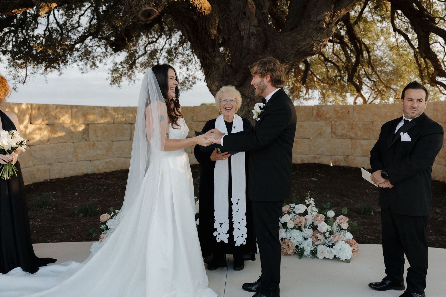 An outdoor wedding ceremony under a tree with floral arrangements and white decor.