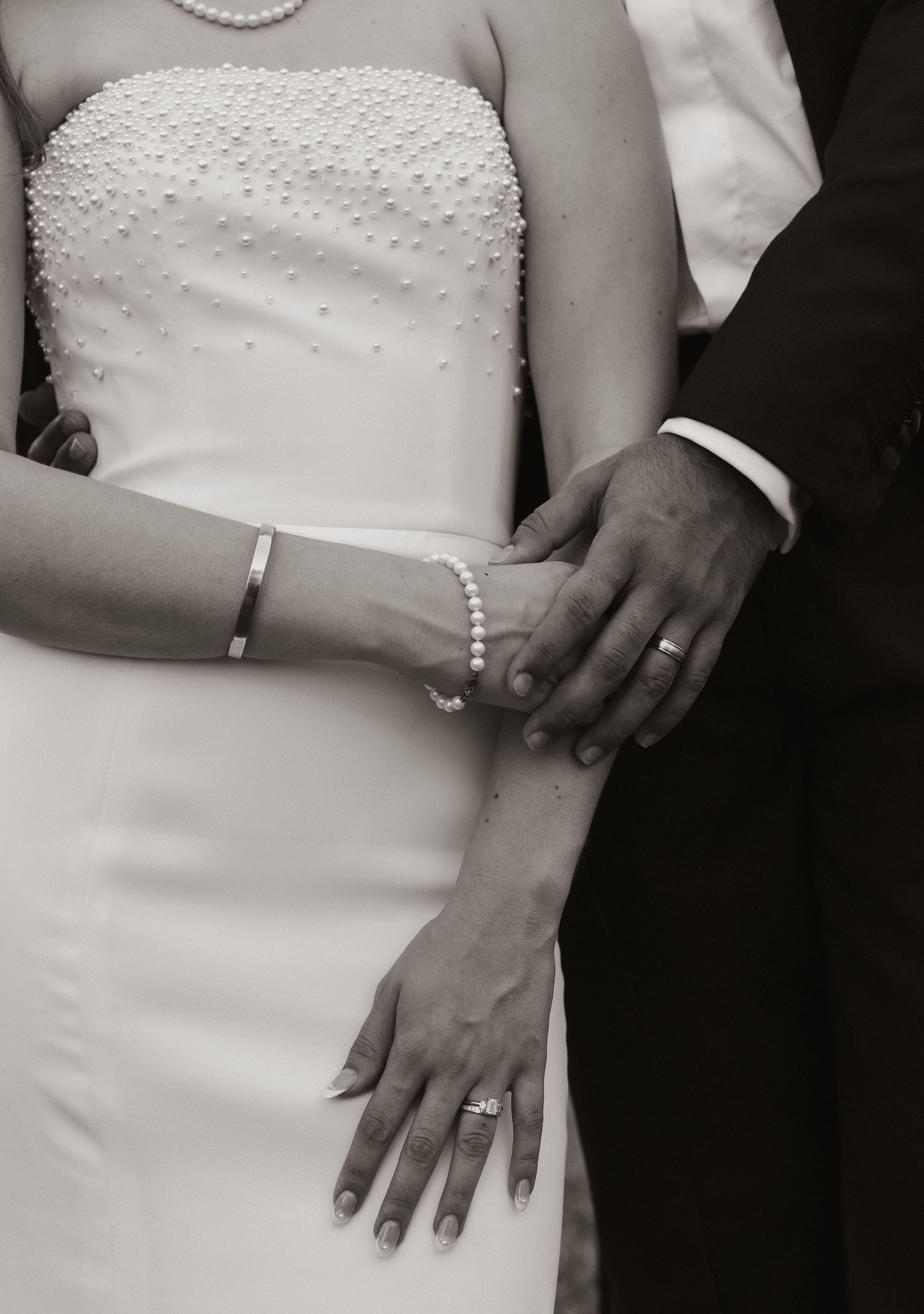 Black and white detail of a bride's beaded wedding dress and groom's hand with rings and pearl bracelet.