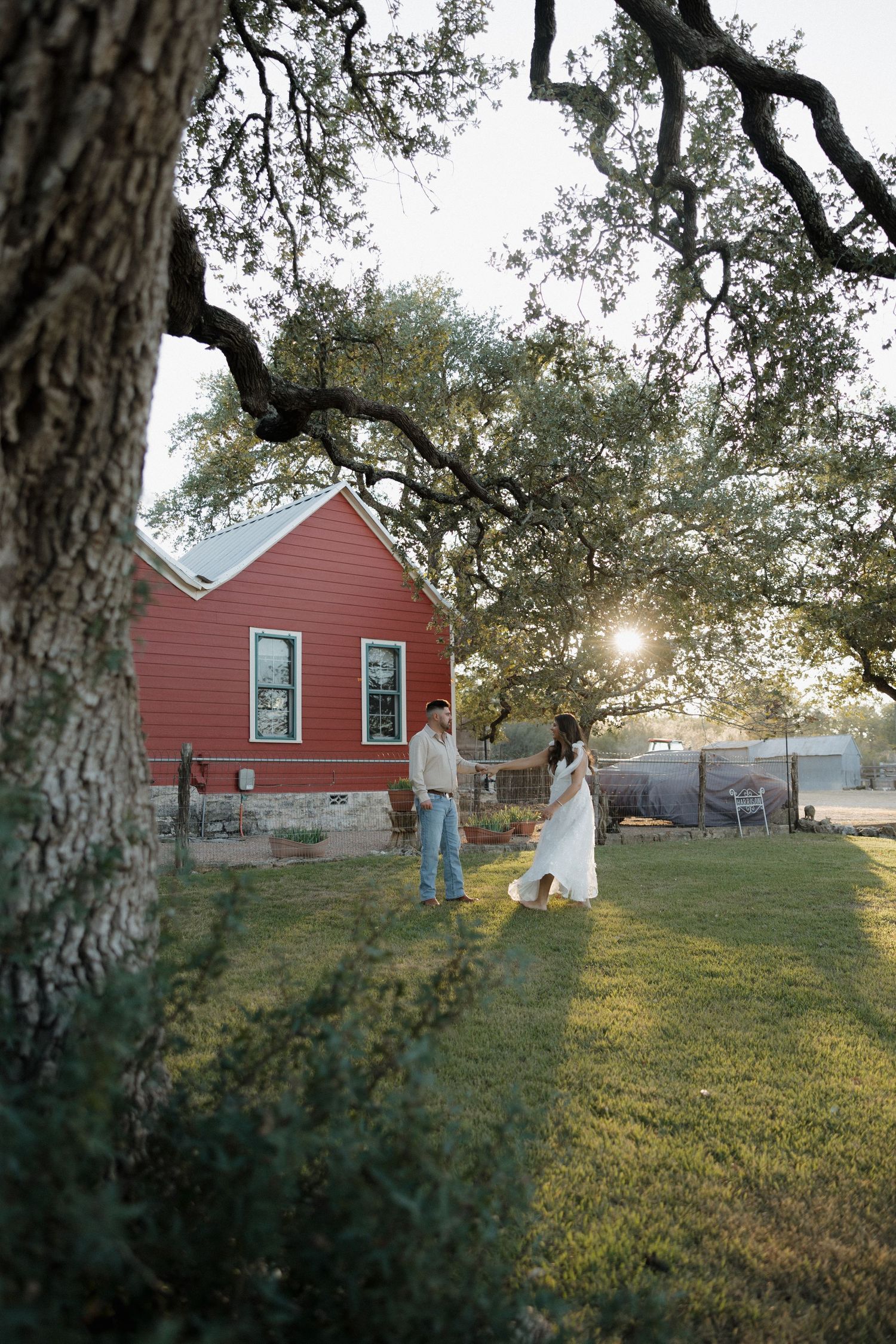A sunset wedding photo near a red house with a large oak tree in the foreground.