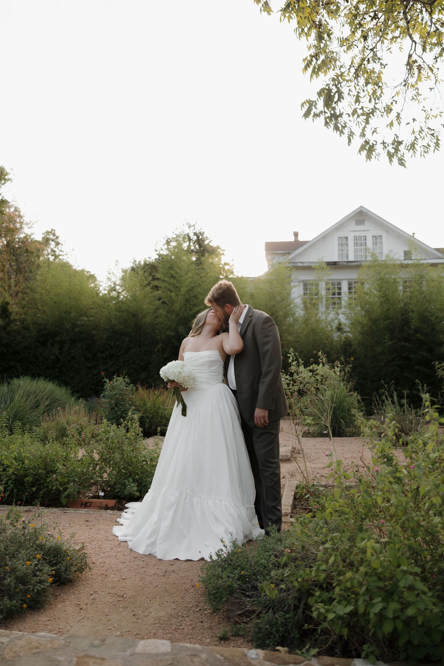 A romantic garden wedding portrait with white house in background during golden hour.