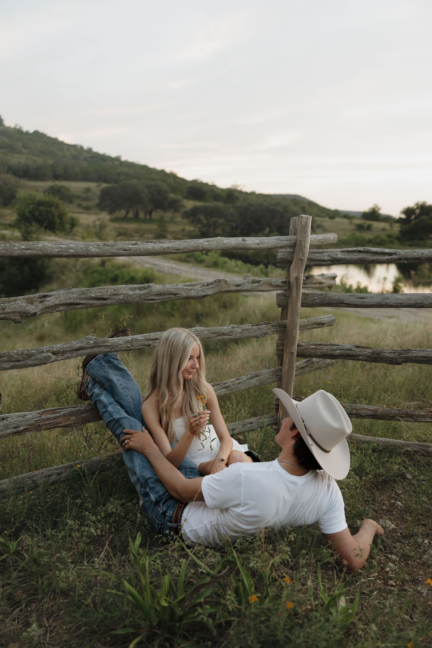 Couple relaxes by rustic wooden fence at sunset near a serene pond in rural countryside setting.