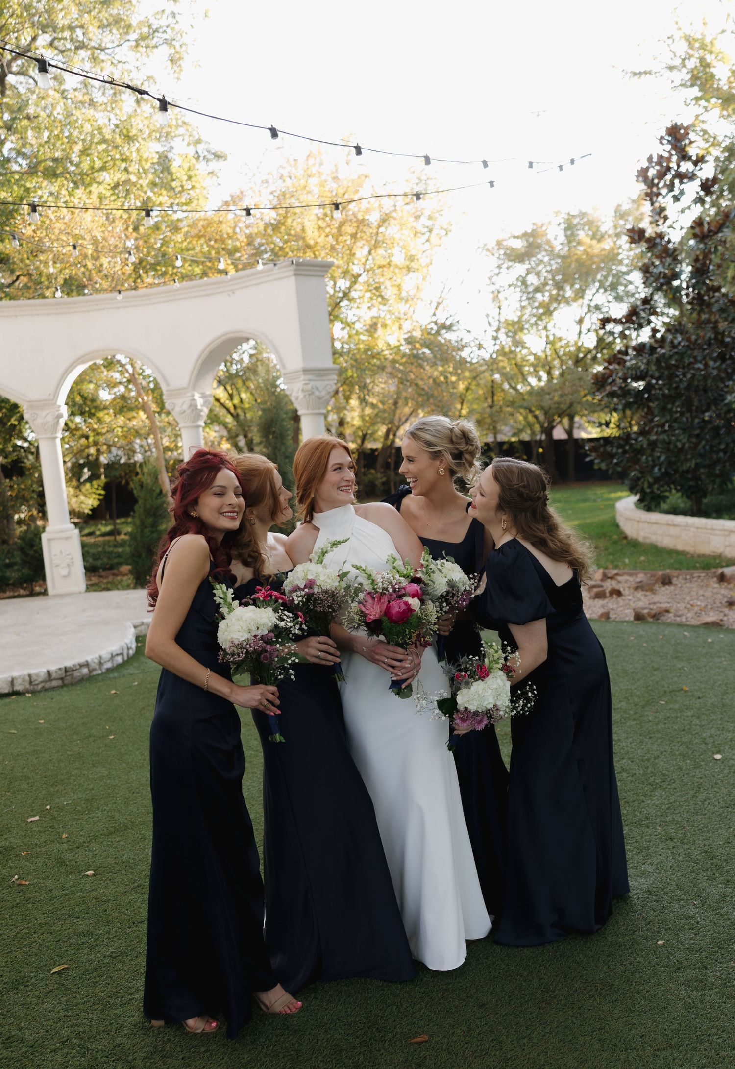 A bridal party in navy blue dresses and white gown gather under string lights at an outdoor venue with arched details.