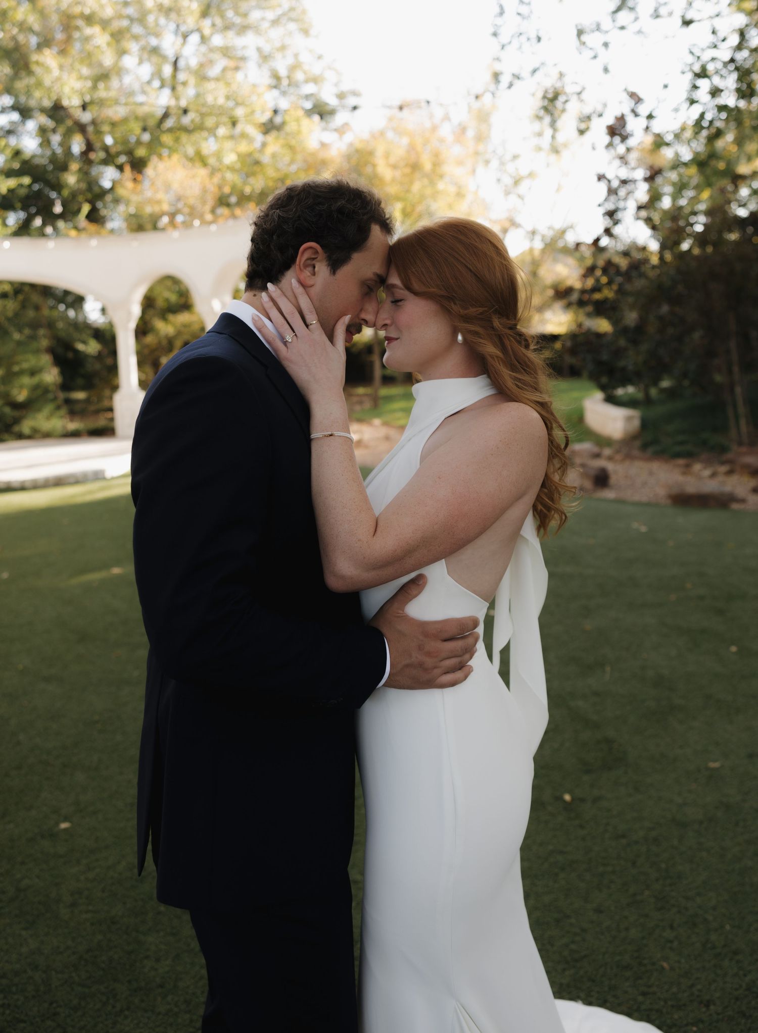 A romantic couple shares a tender kiss in a sunlit garden setting wearing wedding attire.