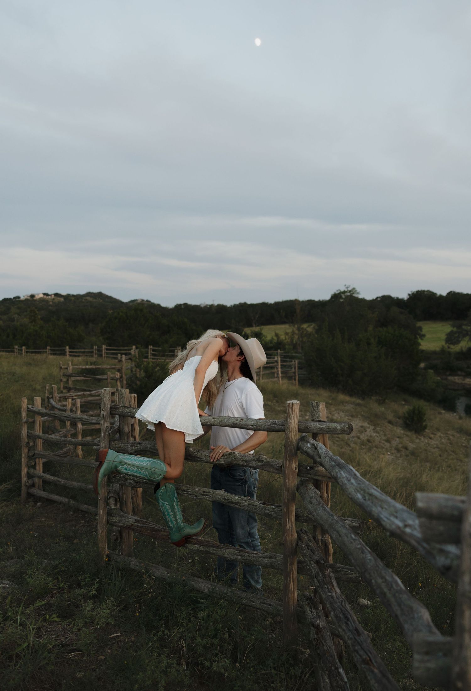 A romantic farm scene with figures sitting on a rustic wooden fence at dusk, with one wearing teal cowboy boots.