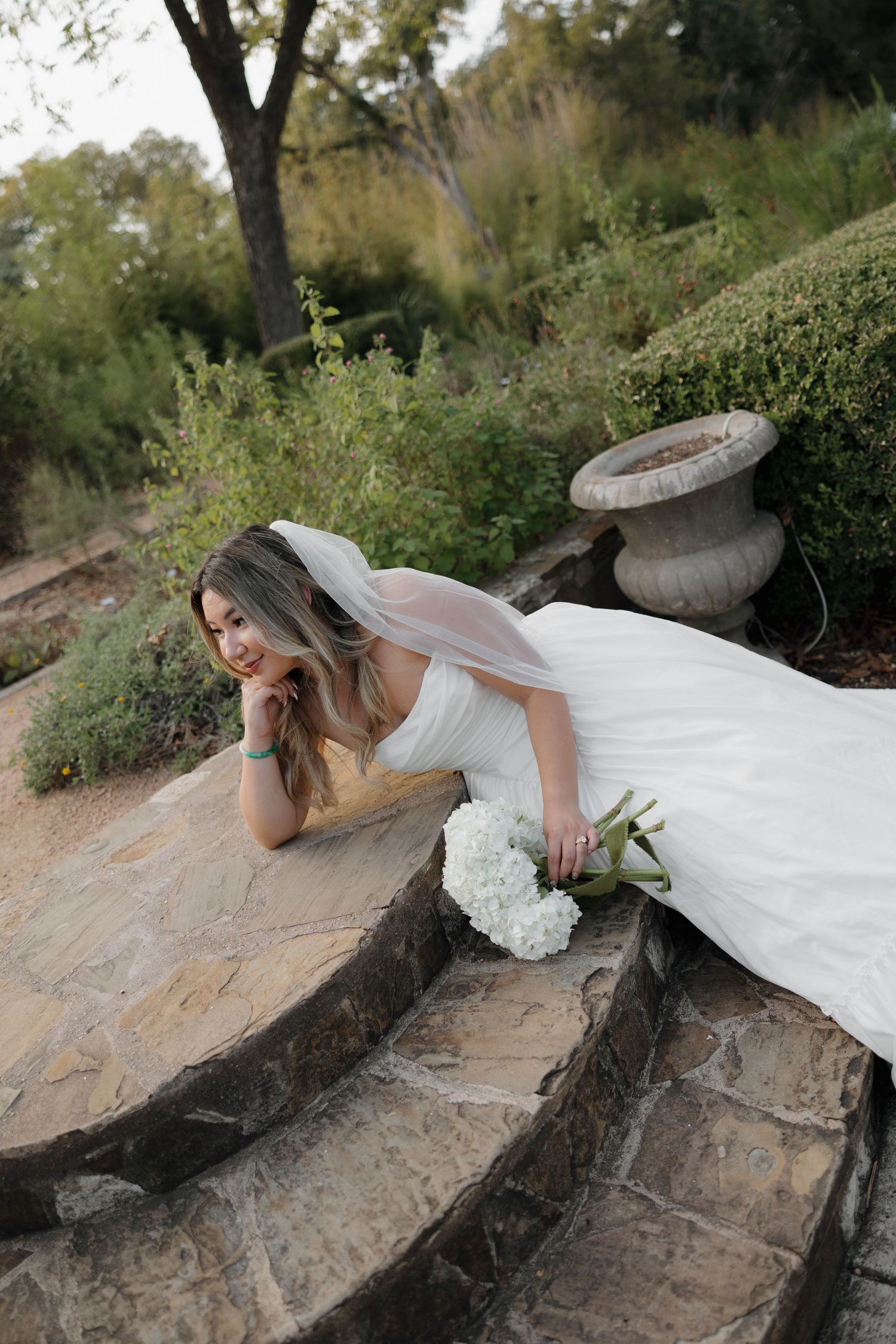 A bride in white dress lounges on stone steps with a bouquet in a garden setting.