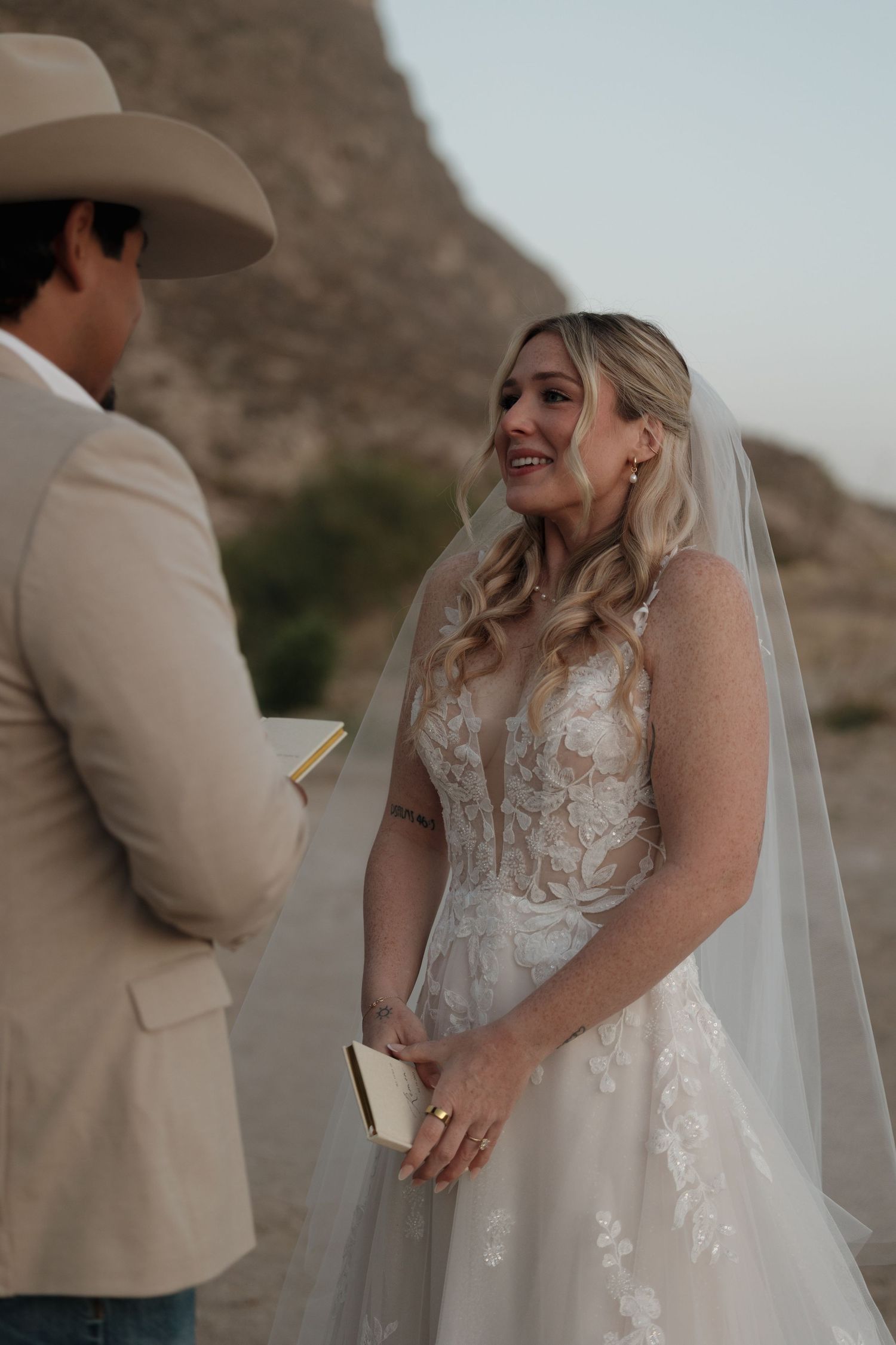 A bride in a lace wedding dress smiles during an outdoor ceremony with mountain backdrop.