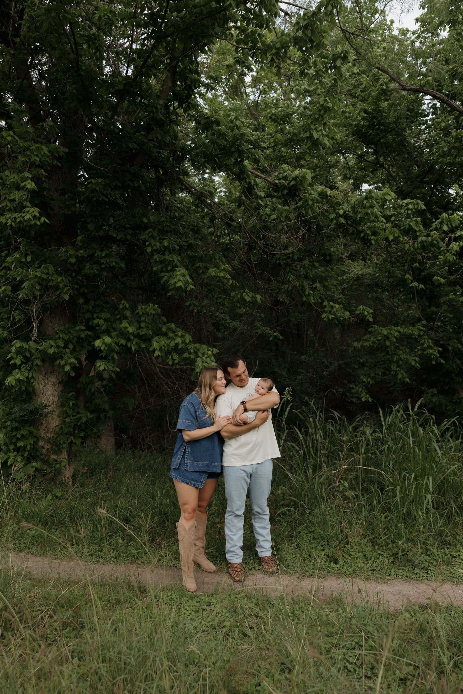 Couple standing together on a dirt path surrounded by lush green trees and tall grass, sharing a tender moment.