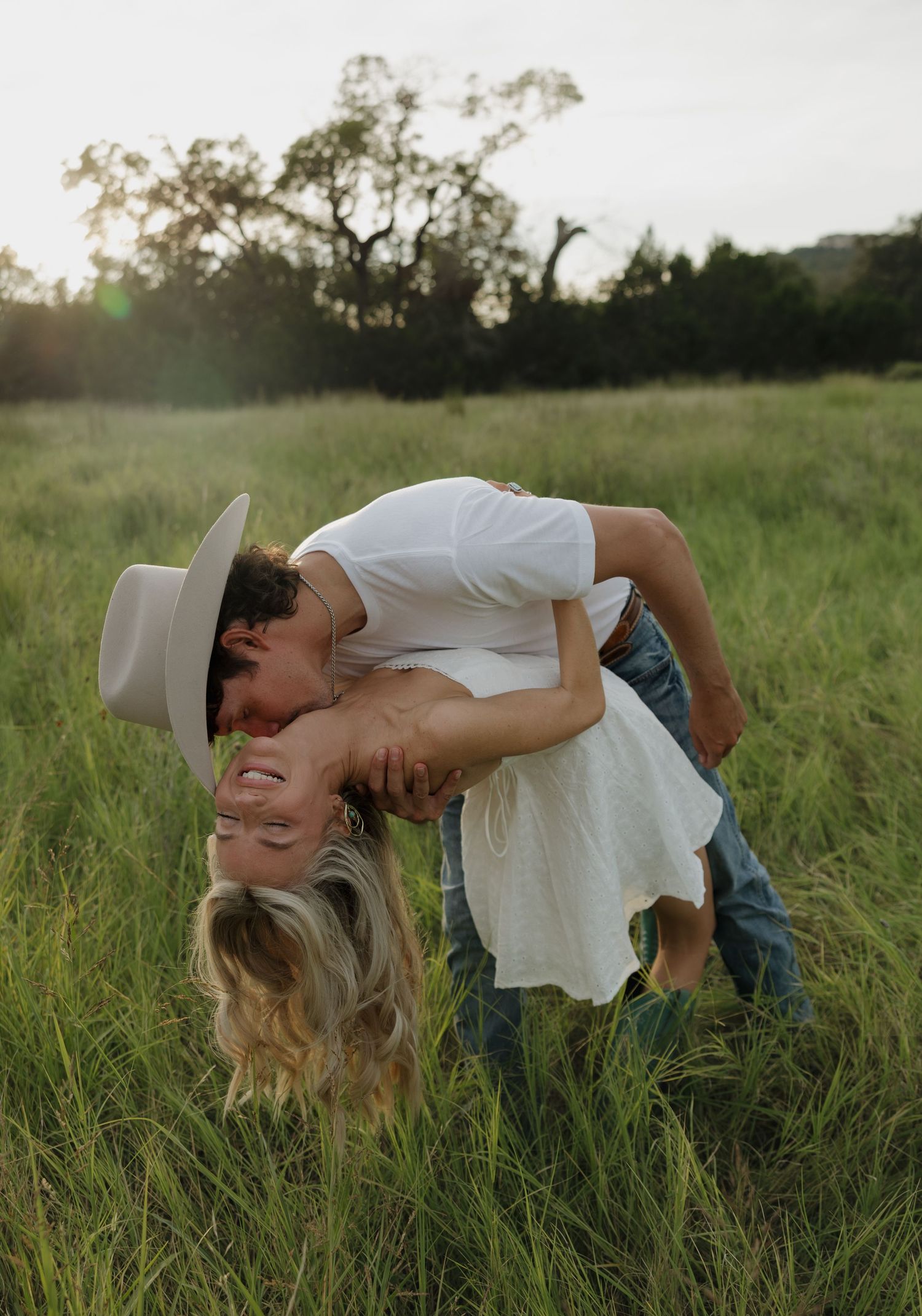 Romantic couple embracing in a sunlit grassy meadow during golden hour with one wearing a white cowboy hat.