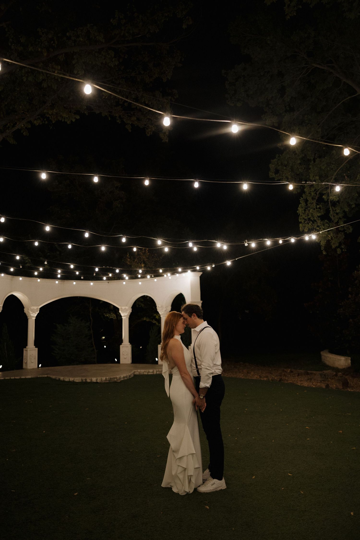 A romantic evening wedding scene with string lights illuminating a couple dancing on a dark lawn near white pillars.