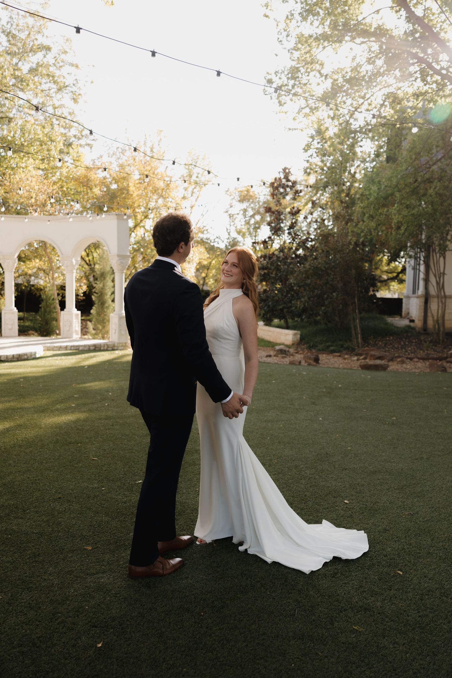 A couple shares an intimate moment on a lawn with string lights and white columns visible in the background.