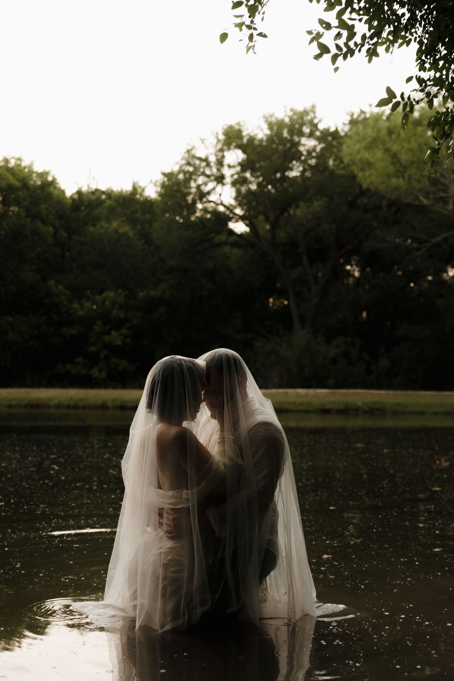 Two figures in wedding veils stand together in a lake during sunset surrounded by trees.