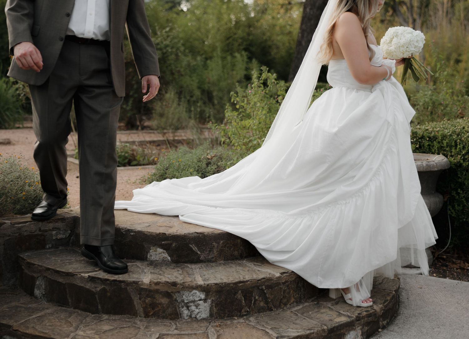 Elegant wedding couple standing on rustic stone steps with flowing white dress swirling in garden setting.