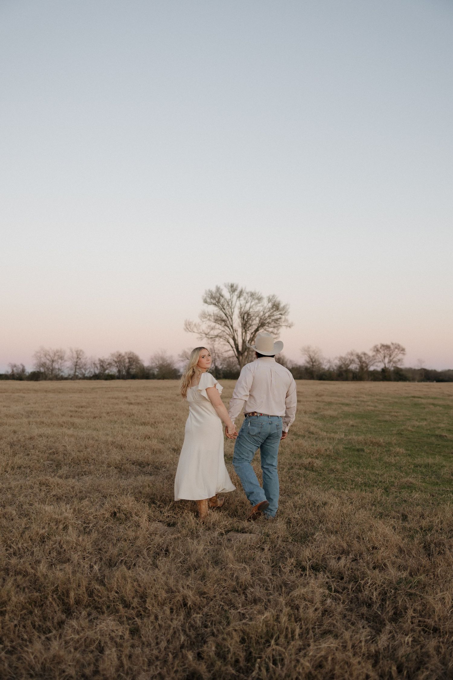 A couple in casual attire walking together in an open field at sunset with a lone tree in the background.