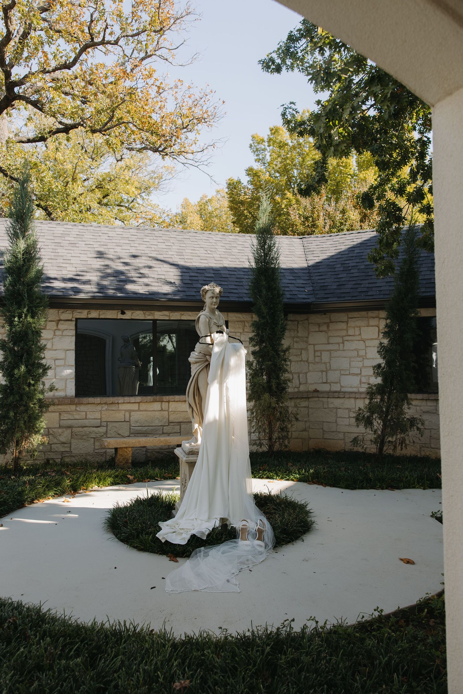 A white wedding dress displayed on a mannequin outdoors on a circular platform against a stone building facade.