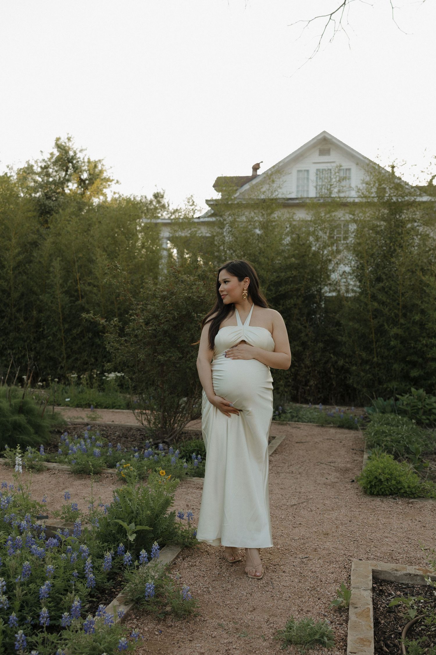 Expectant mother in white dress standing in garden with bluebonnets and white house in background.