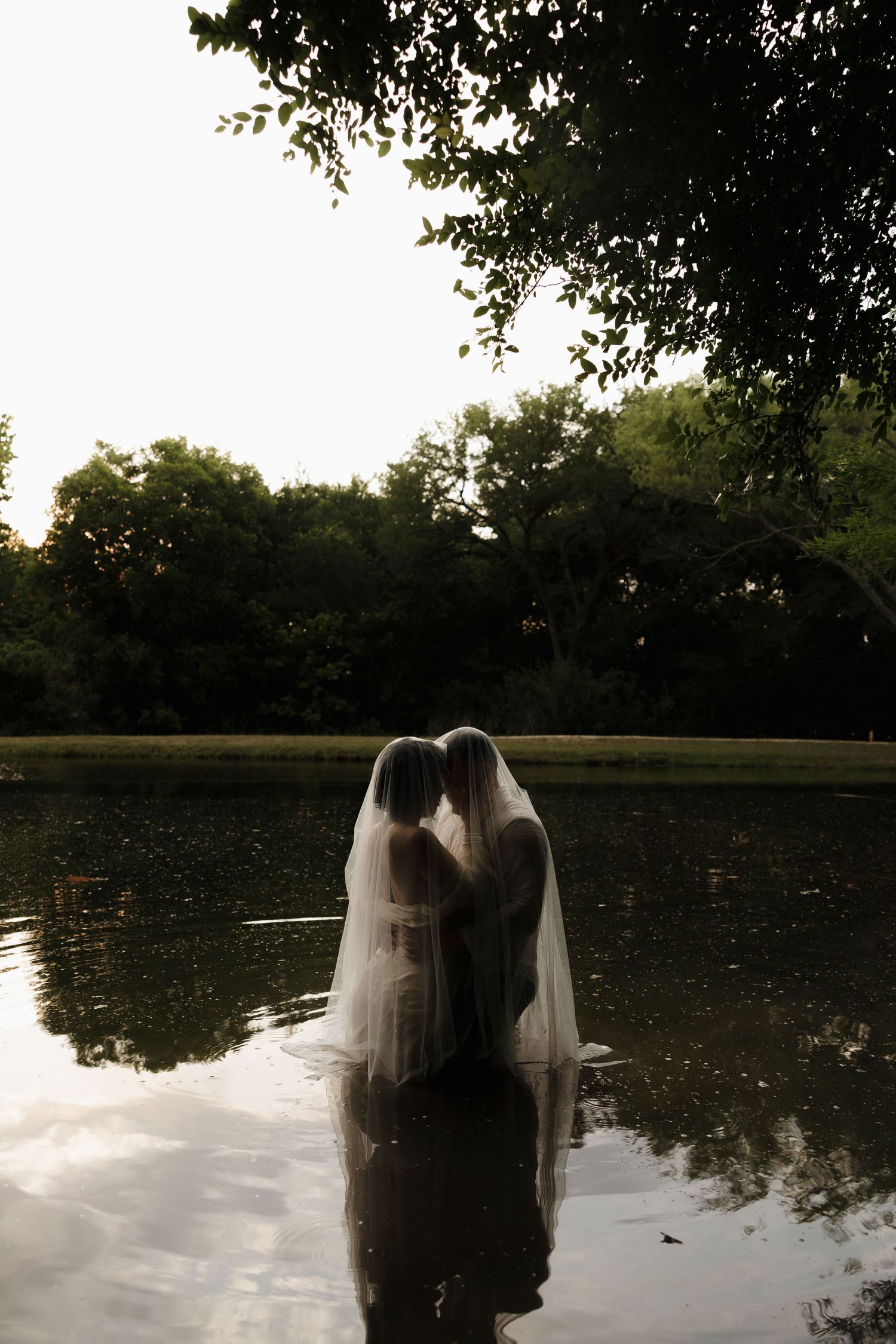 Two figures in white dresses standing in lake with reflection against moody forest backdrop.