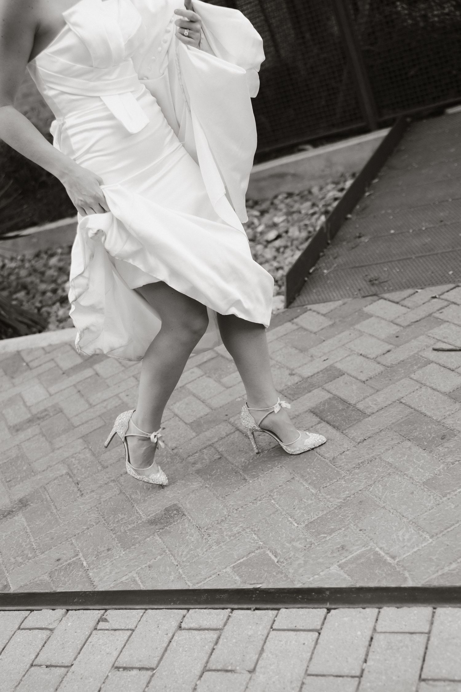 A black and white photo showing feet in white heels and the hem of a flowing white dress on a brick pathway.