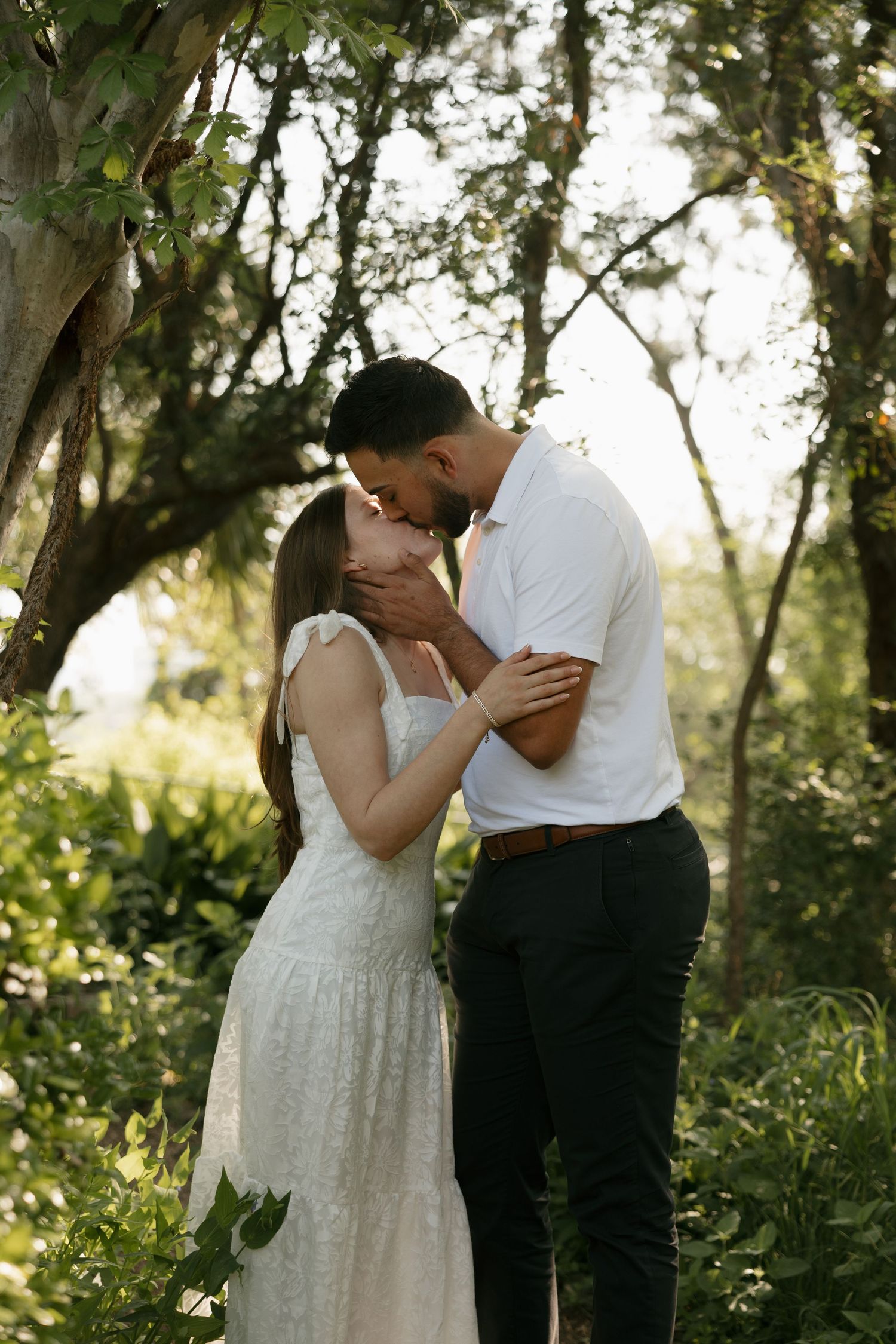 A tender romantic moment captured between a couple in wedding attire surrounded by lush greenery and sunlight.