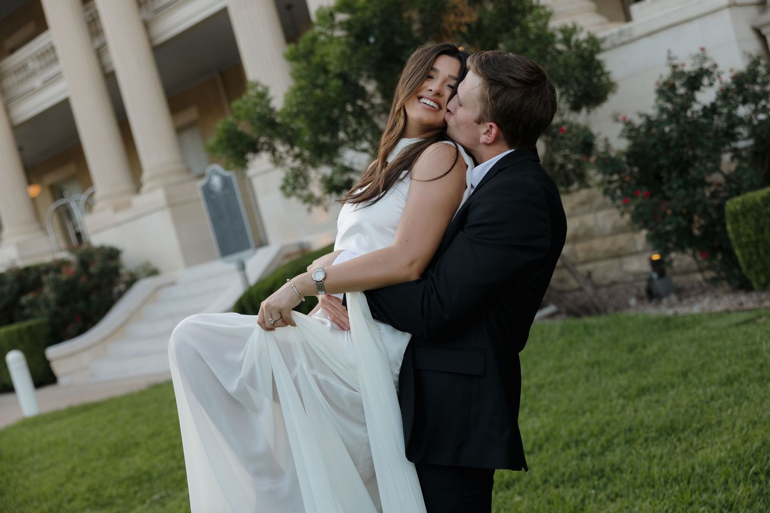 A romantic moment between newlyweds dancing on a grassy lawn near classical architecture.
