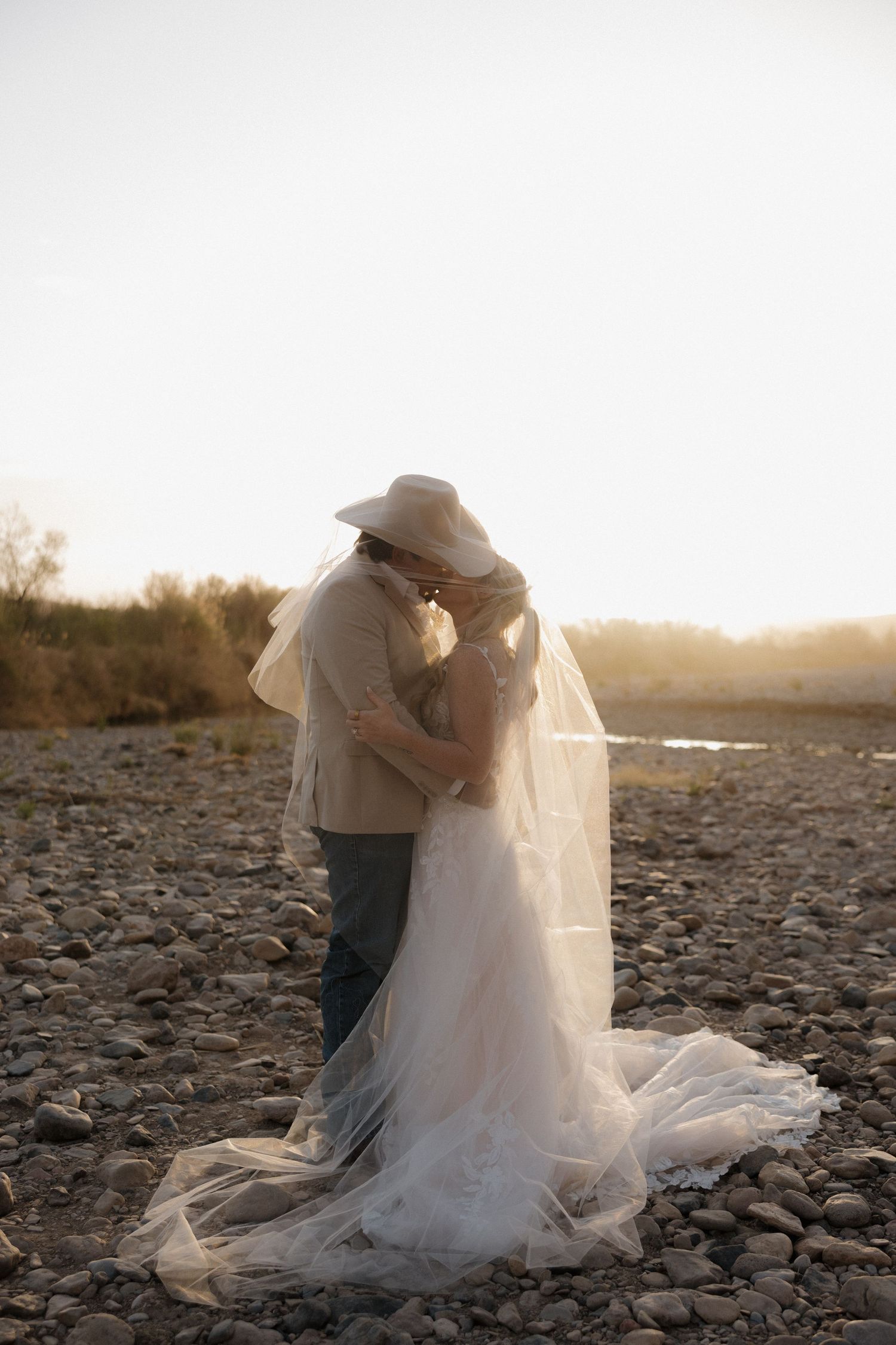 A romantic sunset wedding photo of a couple embracing on rocky terrain with flowing white dress and cowboy hat.