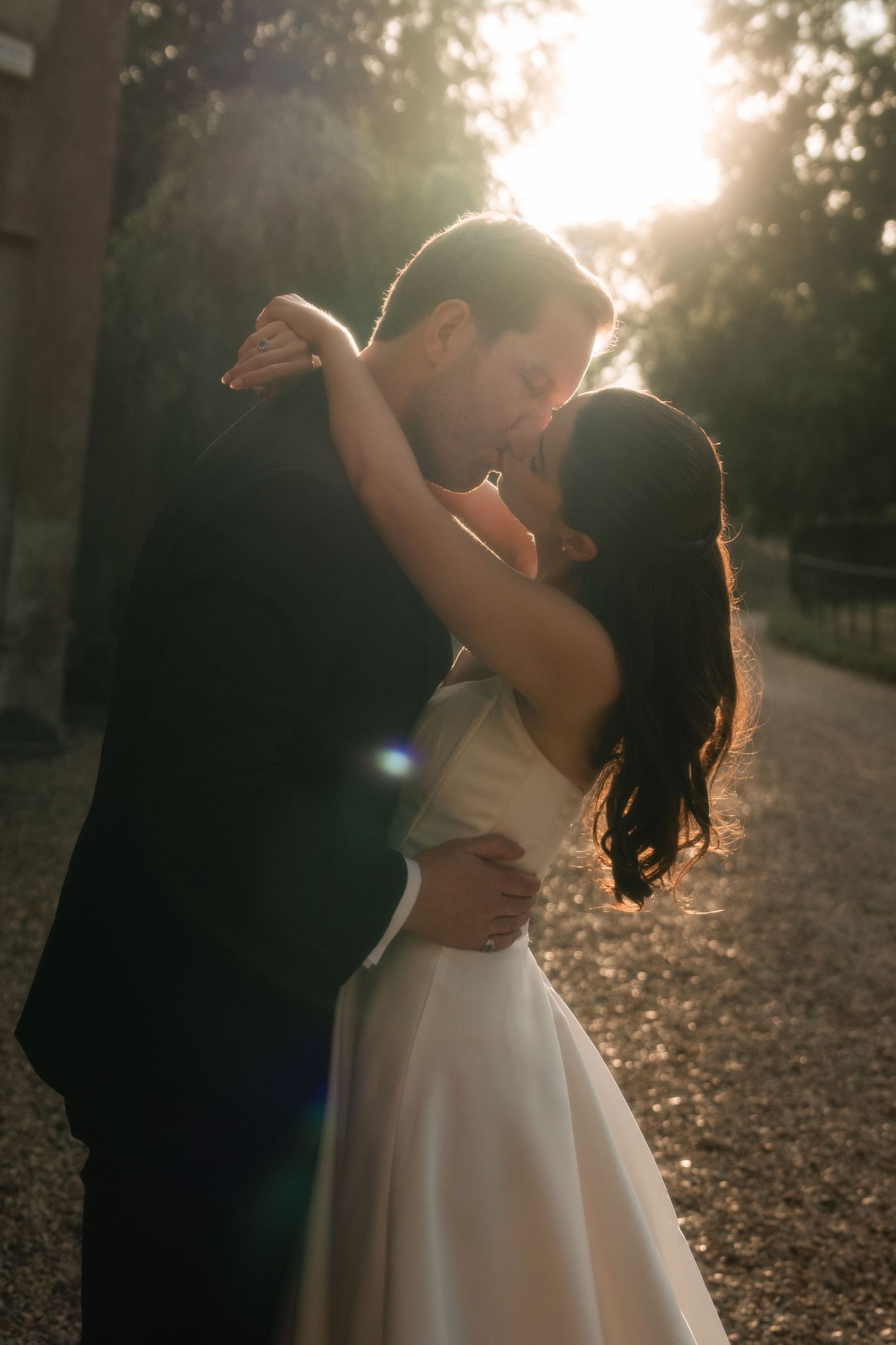 Bride and groom sharing a romantic kiss at golden hour, backlit by warm sunlight in an outdoor setting.