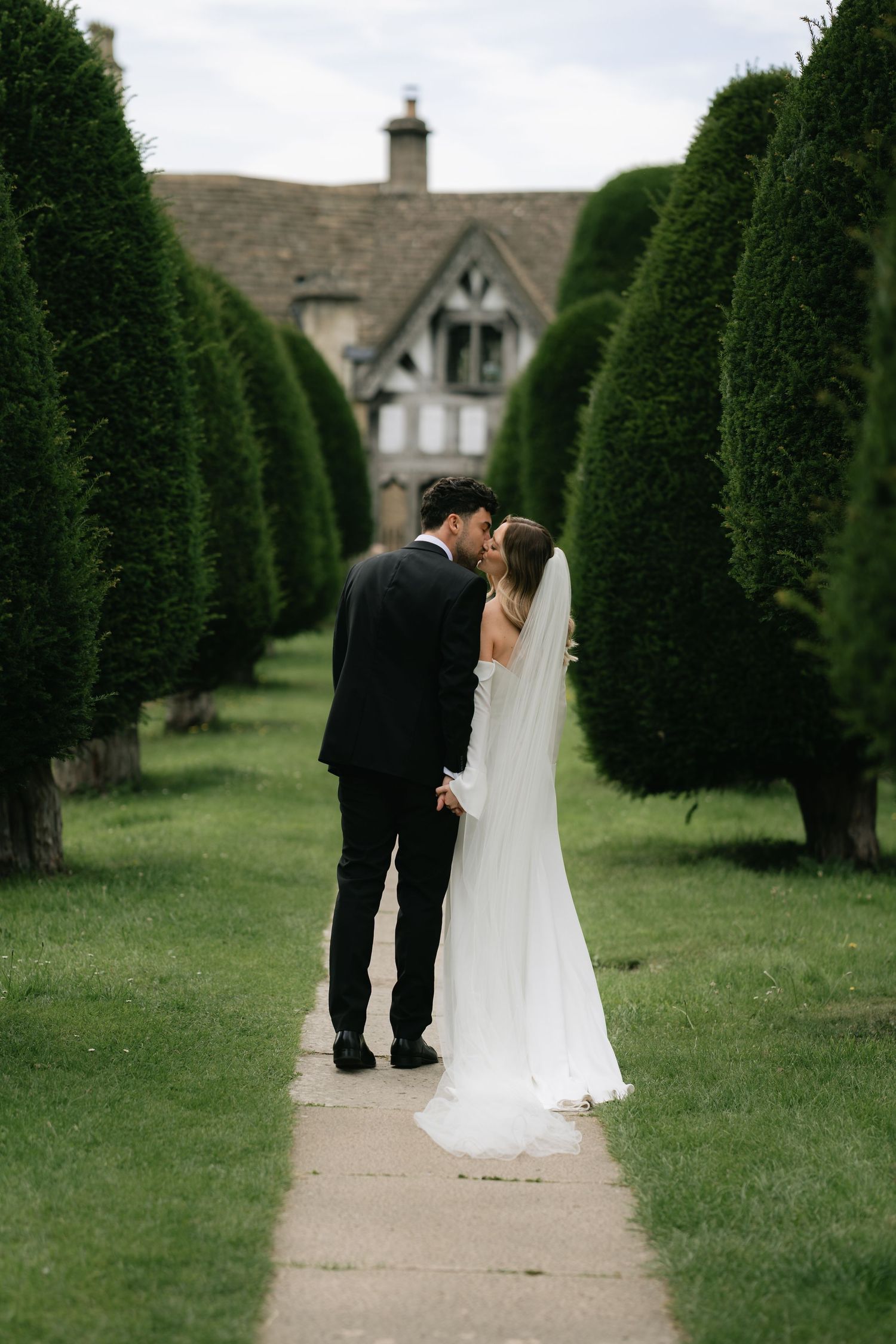 A couple in wedding attire walks along a path lined with perfectly manicured evergreen trees in a formal garden setting.