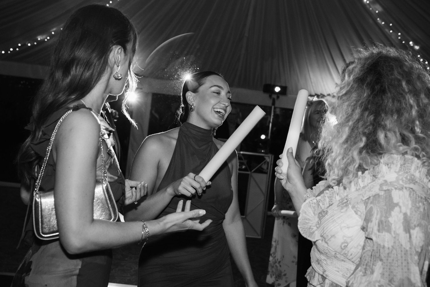 Black and white photo of people dancing and celebrating at a party under string lights in a tent.