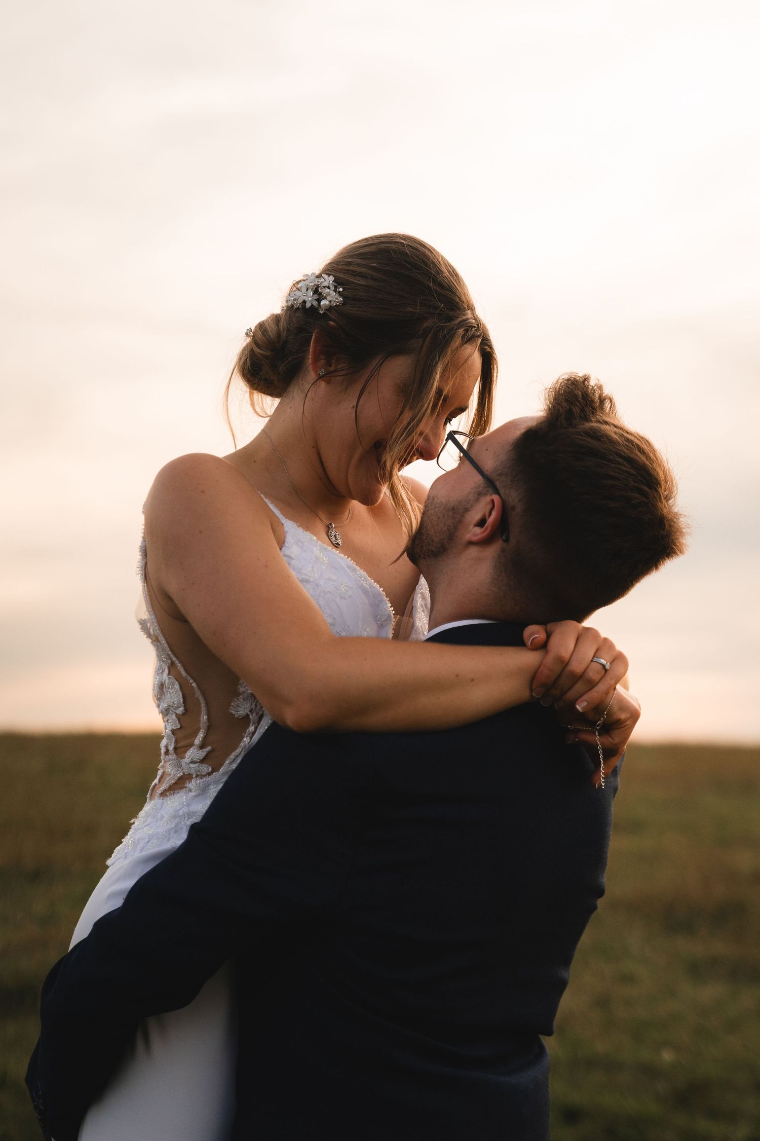 A romantic sunset moment between newlyweds embracing in a field as the bride wears a white lace dress.