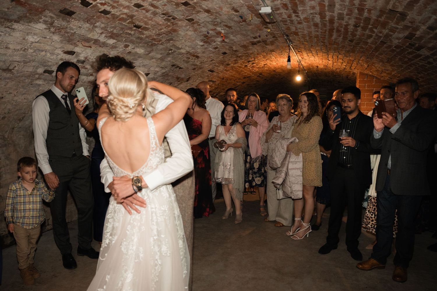 A couple shares their first dance at a wedding reception in a rustic brick-walled venue with guests watching.