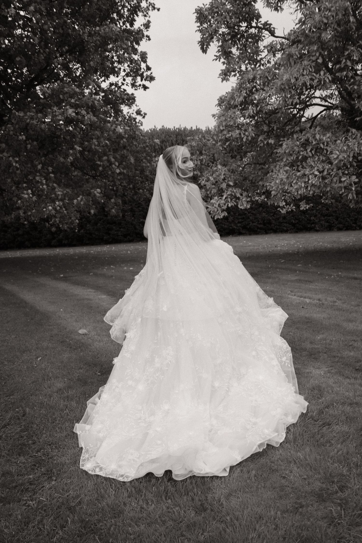 A flowing white wedding dress with a long train spreads gracefully across a grassy lawn in black and white.