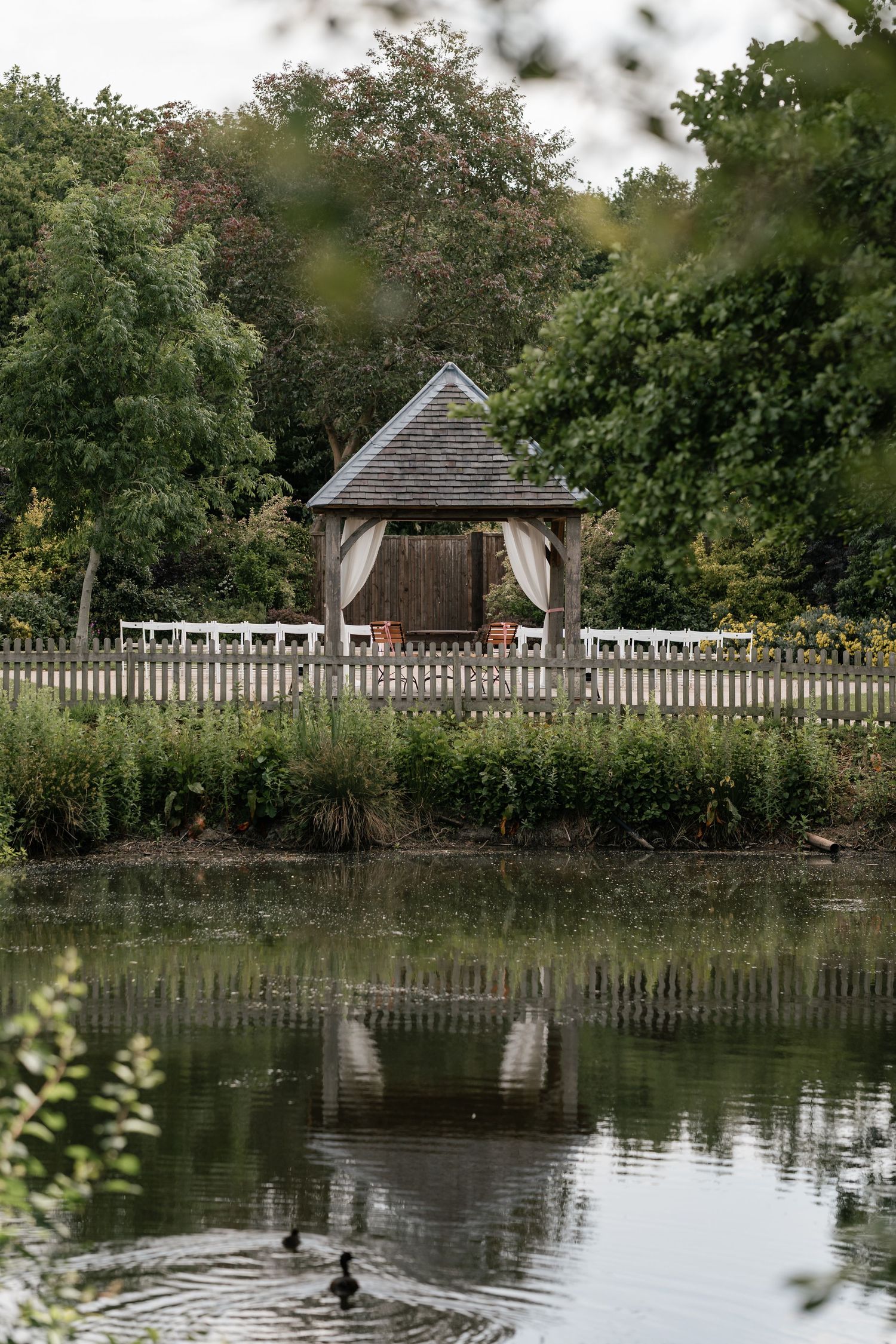 A serene panoramic view of a wooden gazebo and bridge reflected in a tranquil pond surrounded by lush greenery.
