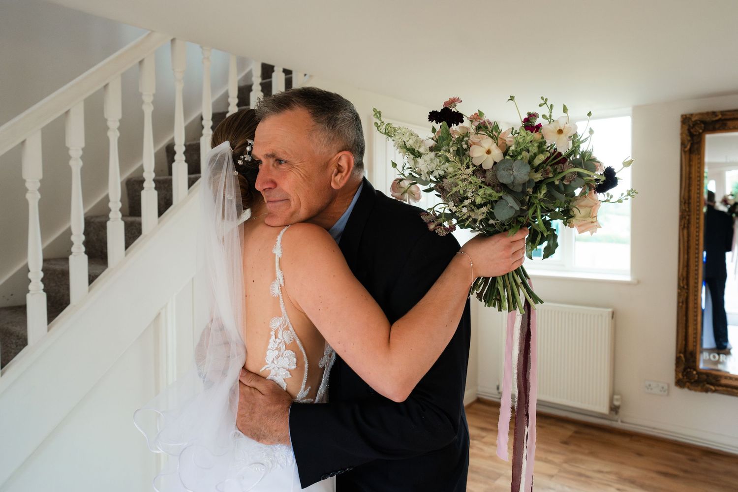 Two people share a tender embrace in front of a white staircase with floral decorations.
