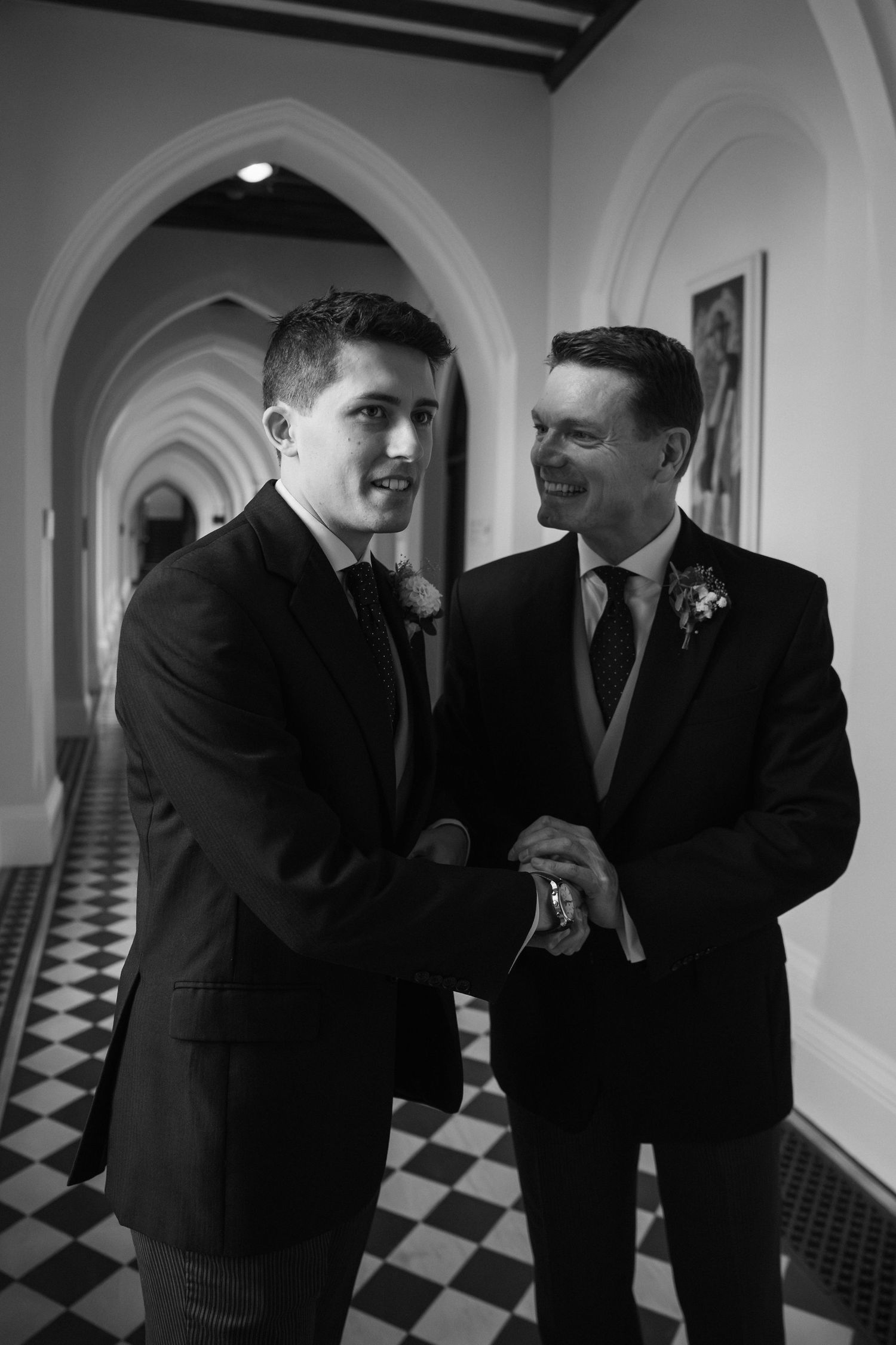 Two people in dark suits share a candid moment in a hallway with black and white checkered floors.