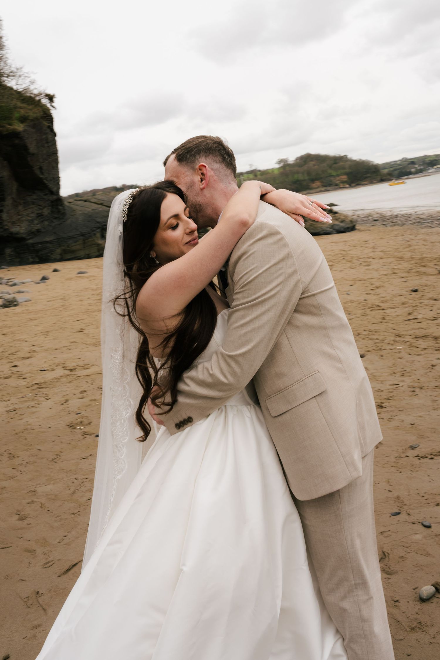 Bride and groom embracing on a sandy beach, bride in white gown with veil, groom in beige suit, overcast sky.